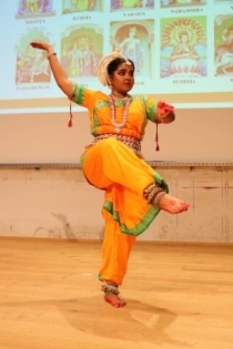 Eine indishe Tänzerin in traditioneller orangener Kleidung mit goldenem Kopfschmuck steht auf einem Bein mit dem anderen angewinkelt in der Luft. Sie hält einen Arm hoch über ihrem Kopf und den anderen vor sich.
