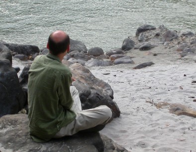 Christian sitzt auf einem Stein am Ufer eines Flusses im Schneidersitz mit dem Rücken zur Kamera und schaut auf das Wasser.